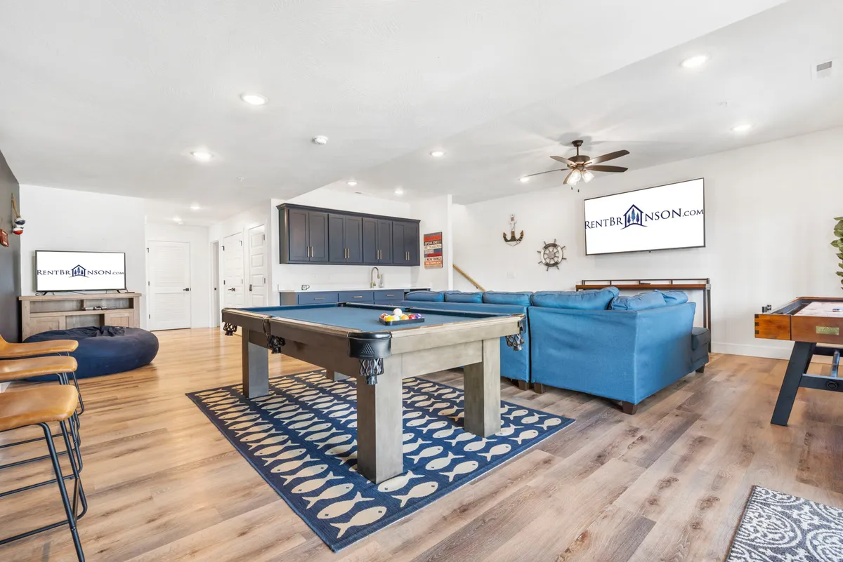A spacious and lively interior game room featuring a pool table, blue sectional sofa, television, and bar stools. The room is designed for fun, relaxation, and group activities, perfect for indoor entertainment during colder or rainy weather.