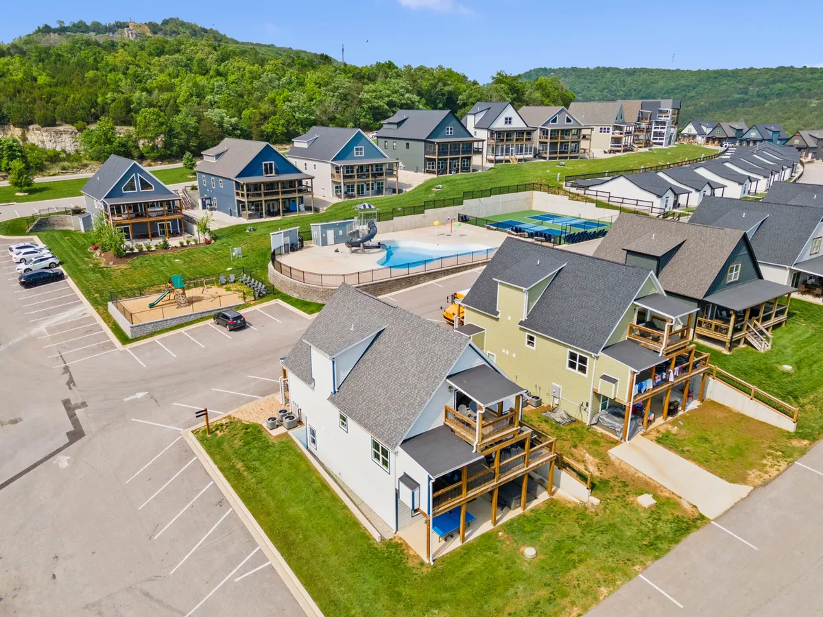 A vibrant exterior view showing a cluster of colorful homes, a large swimming pool area, and an on-site playground nestled against a green hillside. The scene is lively with children playing, families gathering, and a cheerful, community-oriented atmosphere.