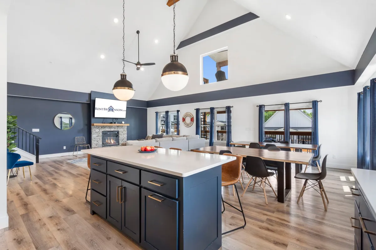 A spacious open-concept living area featuring a large kitchen island, adjacent dining area with wooden tables, a cozy fireplace, and expansive windows that let in abundant natural light. The space is clearly designed for group gatherings, with plenty of seating and an inviting, airy atmosphere.