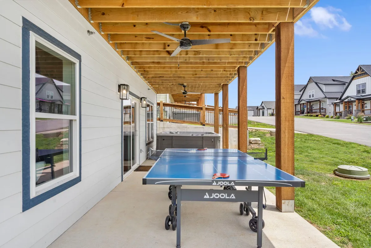 An outdoor patio with a ping pong table, exposed wooden beams above, and a comfortable seating area nearby. The scene evokes a playful outdoor vibe with space for both relaxation and friendly competition.