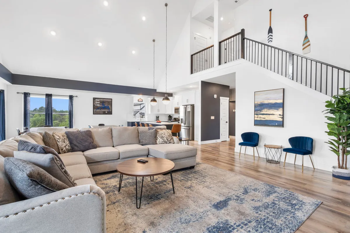 A welcoming living room with a plush sectional sofa circling a round coffee table, decorative wall art, large windows for natural light, and a staircase. The setting is ideal for group relaxation, games, or evening stories by the fire.