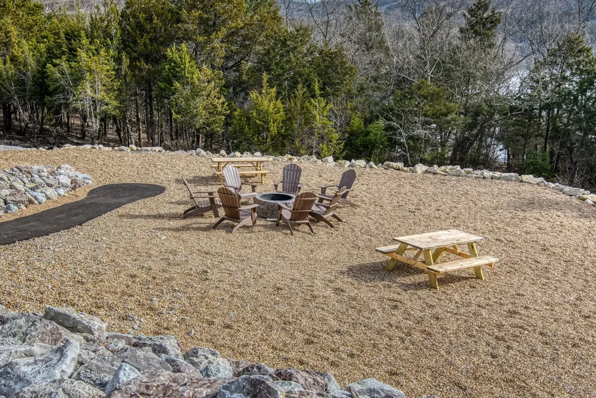 A community fire pit surrounded by Adirondack chairs and a picnic table, nestled near a tree line. The space is set against a serene backdrop, inviting group gatherings and stargazing by the fire.