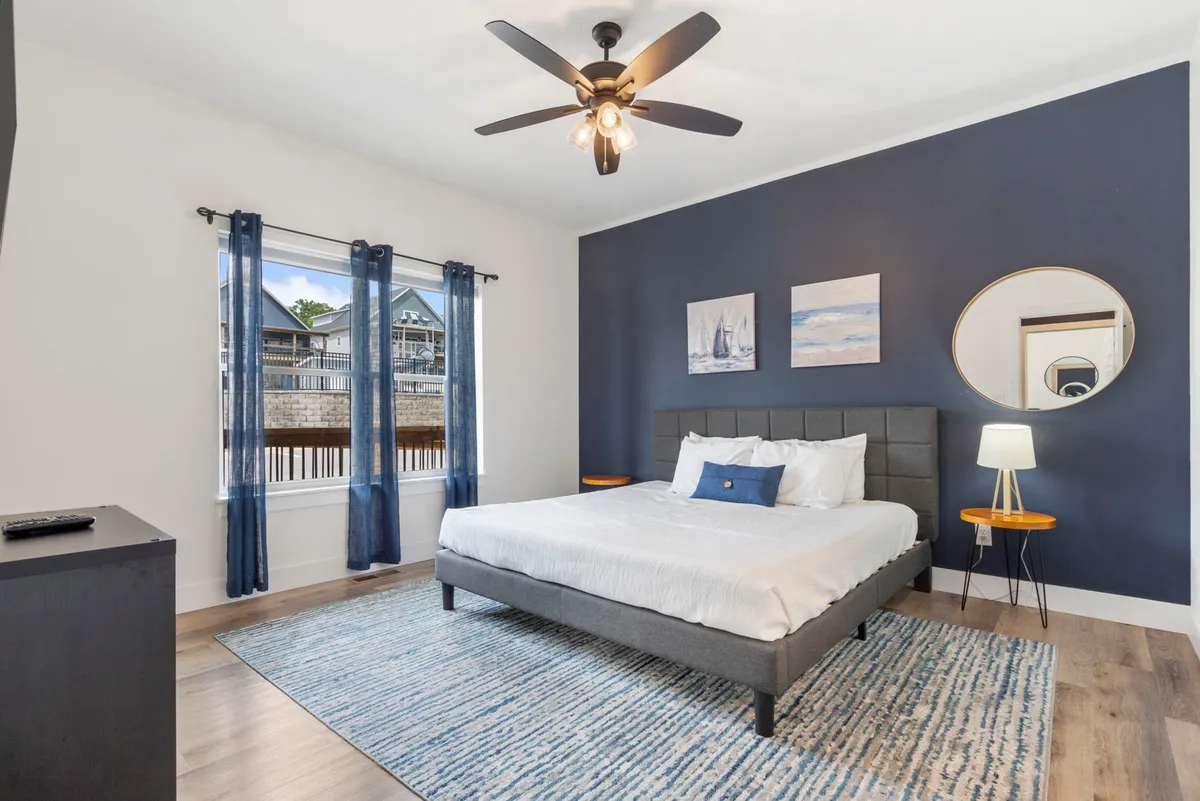 A tranquil bedroom featuring a bed against a blue accent wall, complemented by artwork, a mirror, and a window letting in natural light. The space feels peaceful, perfect for unwinding and enjoying personal downtime.